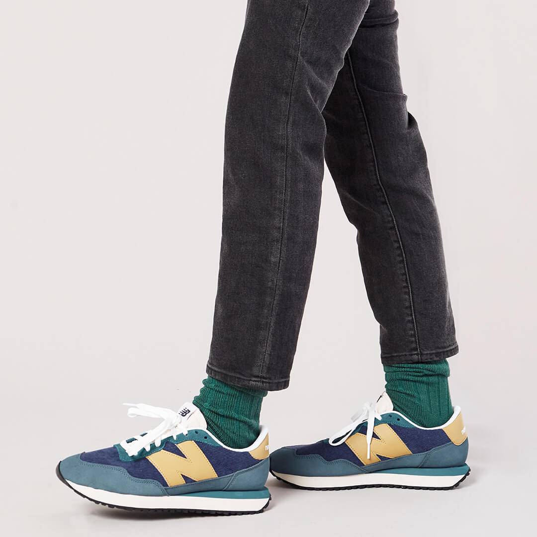 A close-up of a person's feet wearing New Balance 574 sneakers, dark gray jeans, and green crew socks against a light gray studio background.