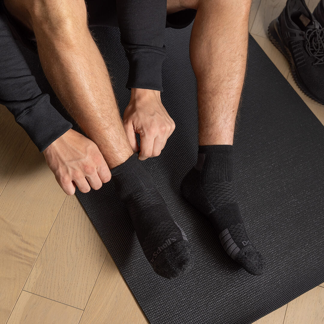 A person sitting on a black yoga mat putting on a black athletic ankle sock, with another black sock already on the foot. A black running shoe is visible on the wooden floor in the background.