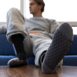 A man reclining on a blue couch with one foot raised, wearing dark gray textured knit socks. The foreground foot is sharply focused, showing the textured sole and toe, while the rest of the scene is softly blurred. thumbnail