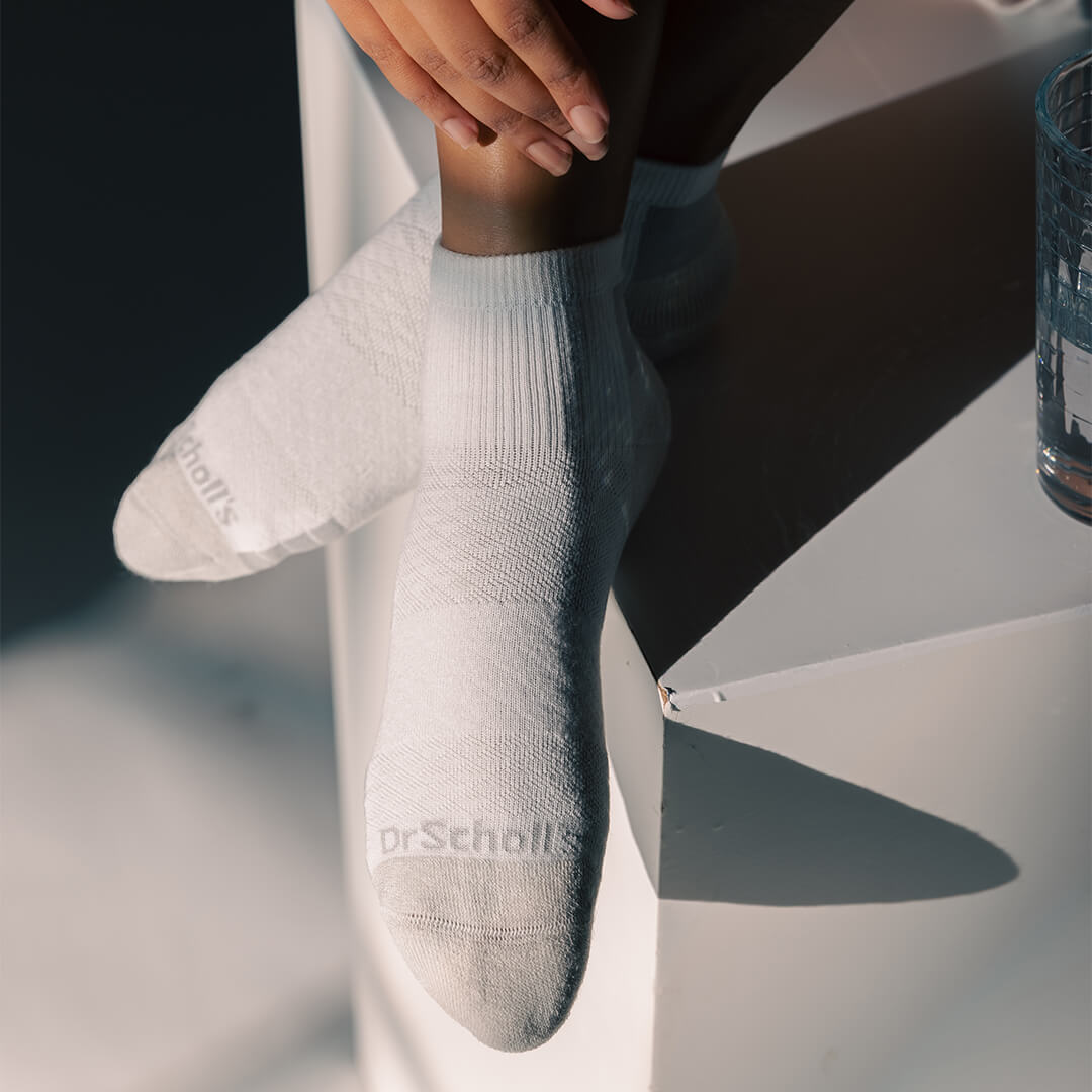 A person wearing white ankle socks with a subtle textured pattern and the brand name 'Dr. Scholl's' printed on the toes, posed on a white geometric surface with dramatic shadows.