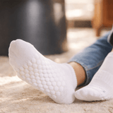 White textured grip socks worn on a person's feet, shown in a cozy indoor setting with blue jeans and a soft beige rug. thumbnail