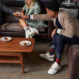 Three people gathered in a lounge setting, with one person reaching across a wooden coffee table to toast with a glass of beer. The person wears a beige jacket, navy pants, white sneakers, and red socks with white socks. thumbnail