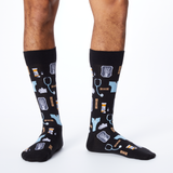 A pair of black crew socks with a medical-themed print of stethoscopes, X-rays, pills, and hands, shown on a model's legs against a white studio background. thumbnail