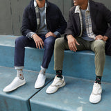 Two men sitting on blue concrete steps wearing white low-top sneakers and HOTSOX sports-themed novelty crew socks. They are dressed in casual jackets, shirts, and jeans or chinos.
