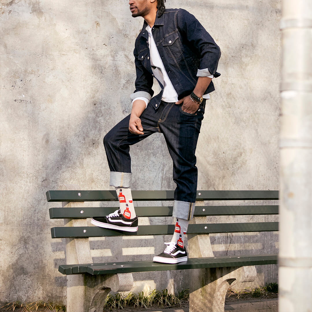 A man wearing a dark denim jacket, dark jeans, a white t-shirt, and black Vans sneakers with red and white socks, posing on a green wooden bench against a textured concrete wall.