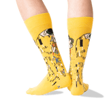 A pair of bright yellow crew socks with a Gustav Klimt-style art print featuring black, white, green, and orange details, shown on a person's legs against a clean white background.