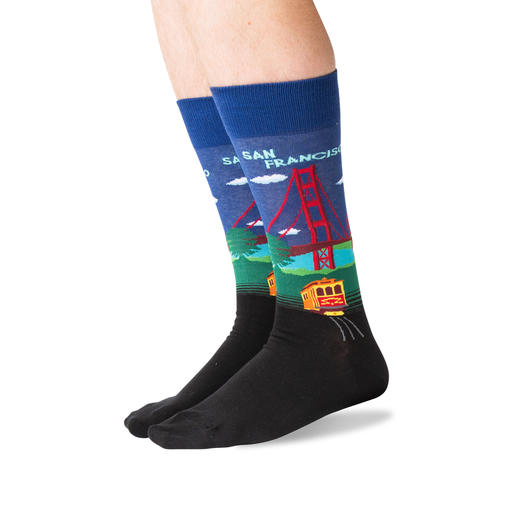 A pair of crew socks worn on a person's feet, featuring a colorful graphic of the Golden Gate Bridge and a trolley train in San Francisco on a blue background, with a black toe and heel section.
