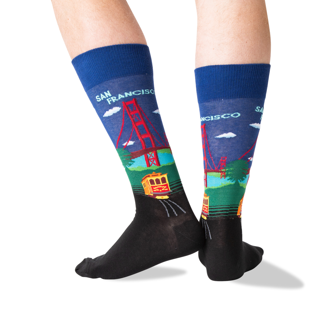 A pair of crew socks worn on feet, featuring a colorful San Francisco-themed graphic with the Golden Gate Bridge, trolleybus, and 'SAN FRANCISCO' text on a white background.