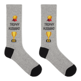 A pair of heather gray crew socks with black heels and toes, featuring a gold trophy graphic and the text 'TROPHY HUSBAND' printed on the front.