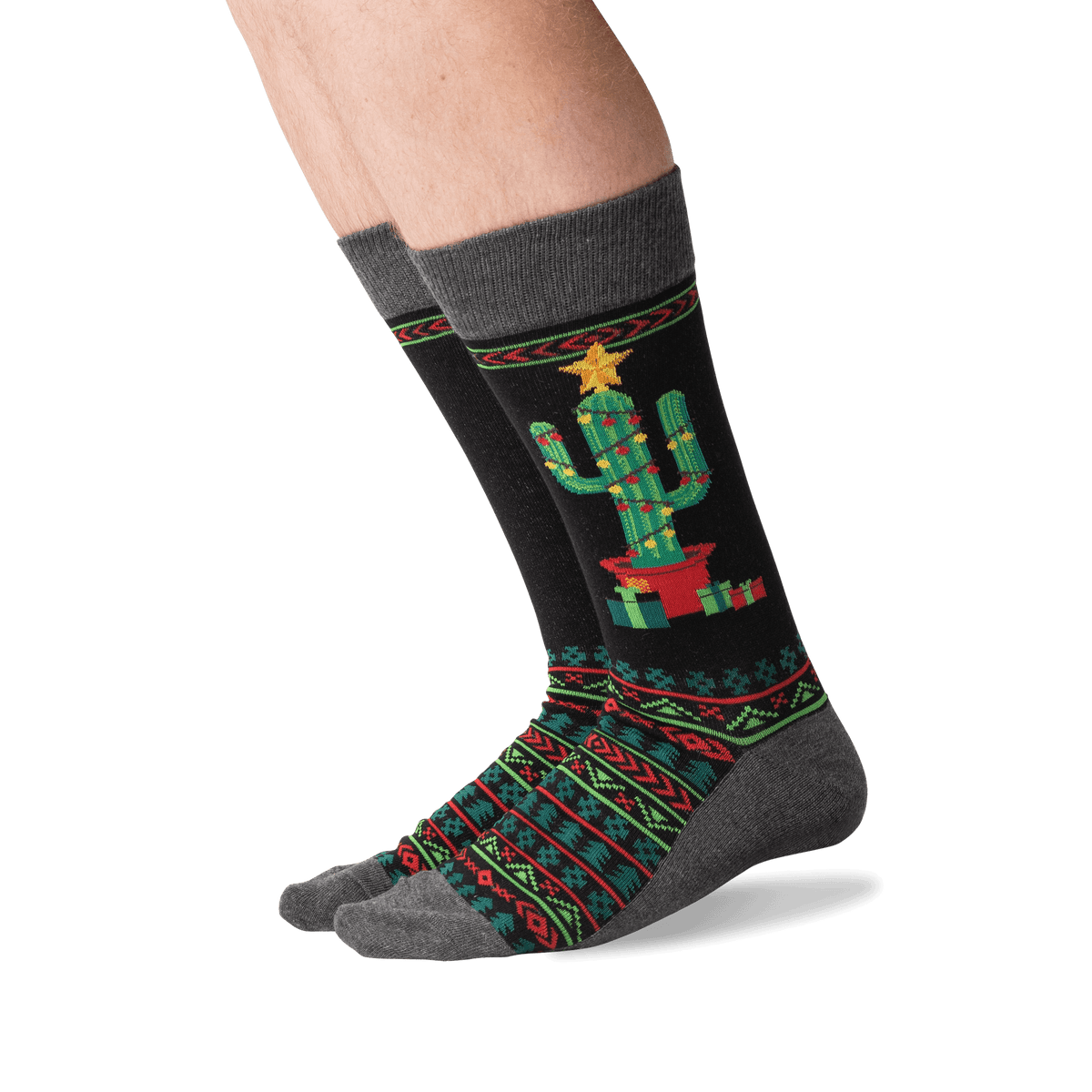 A pair of crew socks worn on a person's feet, featuring a black background with a green cactus in a red pot, wrapped in festive red and green geometric patterns, and topped with a yellow star.