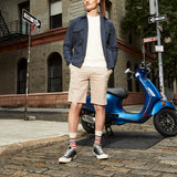 A man stands on a city street wearing a navy overshirt, cream T-shirt, beige shorts, striped socks, and black-and-white sneakers, with a blue scooter and brick buildings in the background. thumbnail