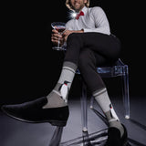 A man sits on a transparent chair wearing gray socks with penguin illustrations, black loafers, a white dress shirt, black trousers, and a red bow tie, while holding a martini glass against a black background.