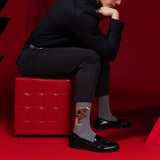 Person sitting on a red stool wearing black shoes and gray socks with The Thinker design, against a red background. thumbnail