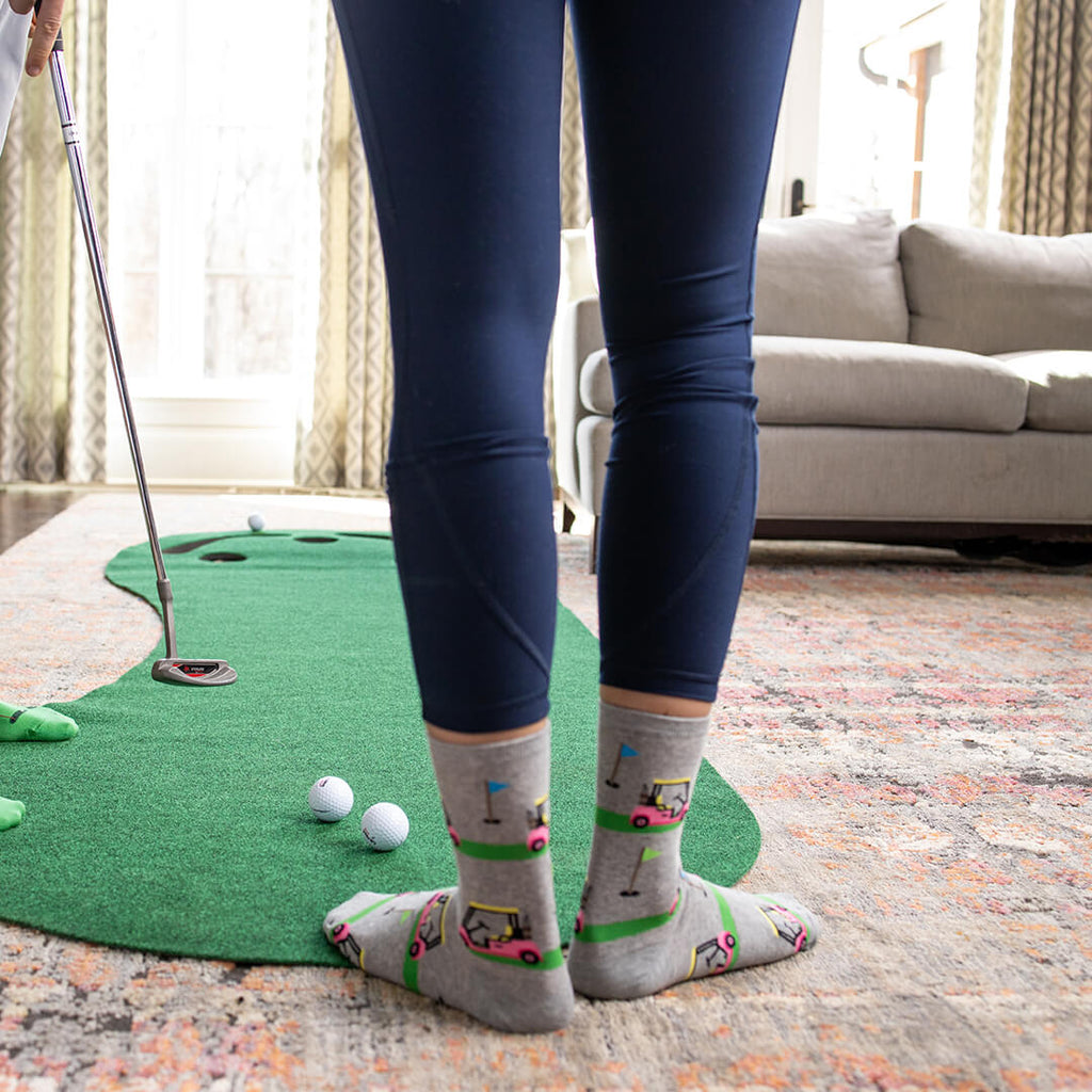 Person playing mini golf indoors with a green mat and golf ball on a patterned rug.