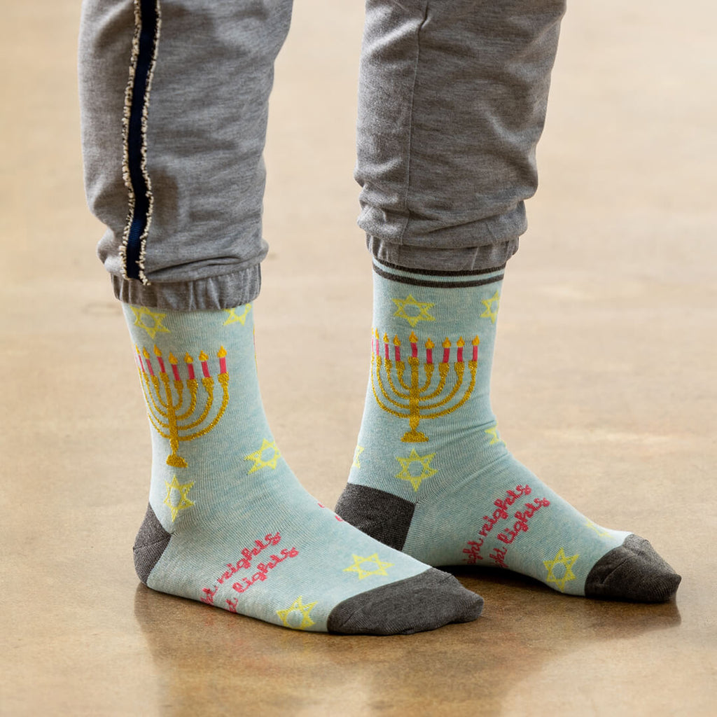 A person wearing light blue crew socks with a yellow menorah, pink candles, and pink text, paired with gray sweatpants, standing on a wooden floor.