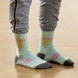 A person wearing light blue crew socks with a yellow menorah, pink candles, and pink text, paired with gray sweatpants, standing on a wooden floor. thumbnail