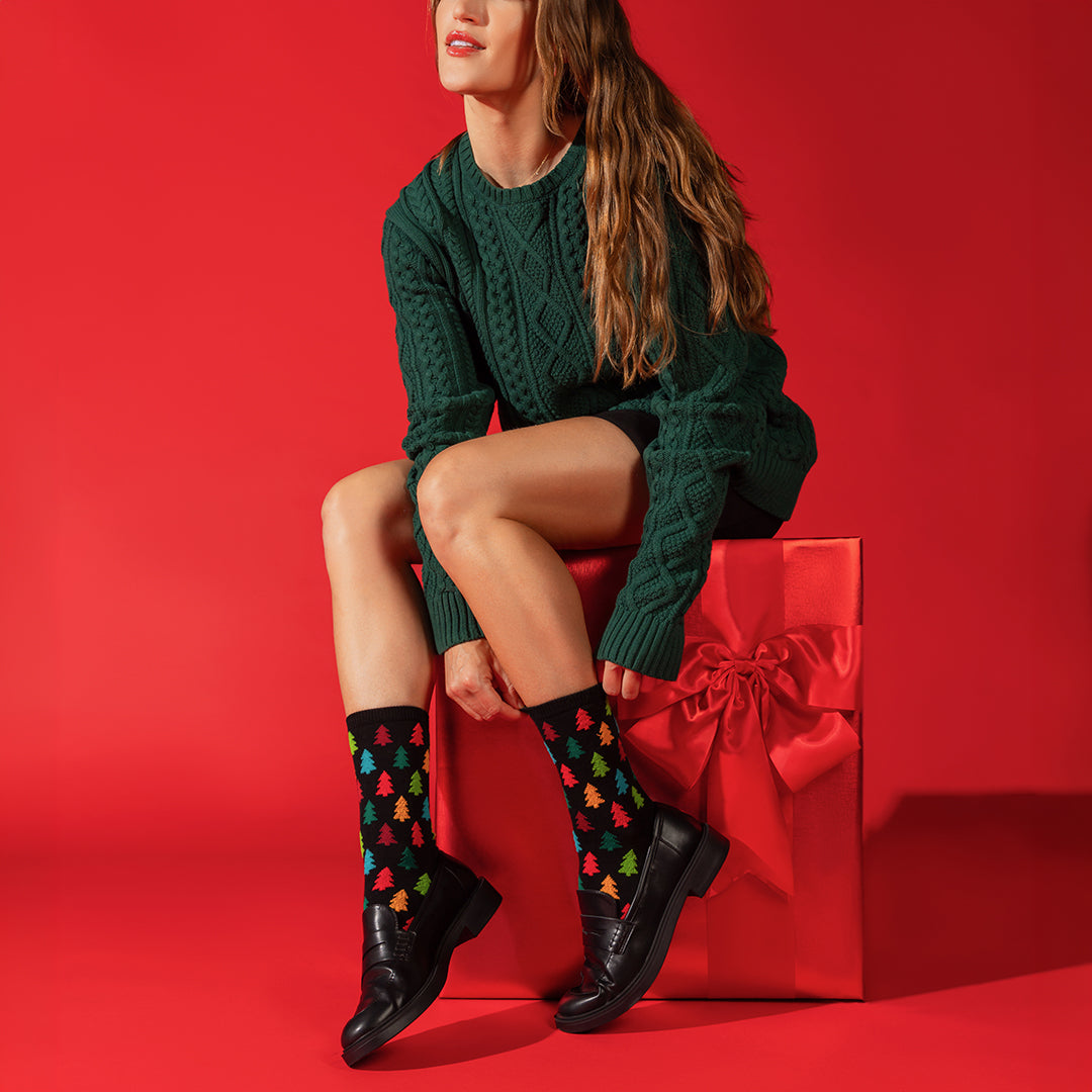 A woman sits on a large red gift box against a solid red studio background, wearing a dark green cable-knit sweater dress, black leather shoes, and black crew socks with a colorful Christmas tree pattern.