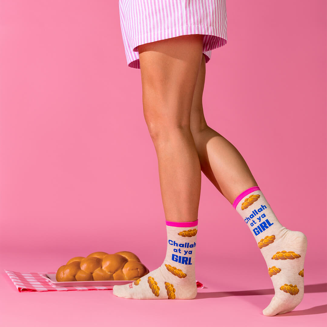 A person wearing cream crew socks with pink cuffs and blue text that read 'Challah at ya GIRL' stands against a pink studio background. The socks also have a small challah print.