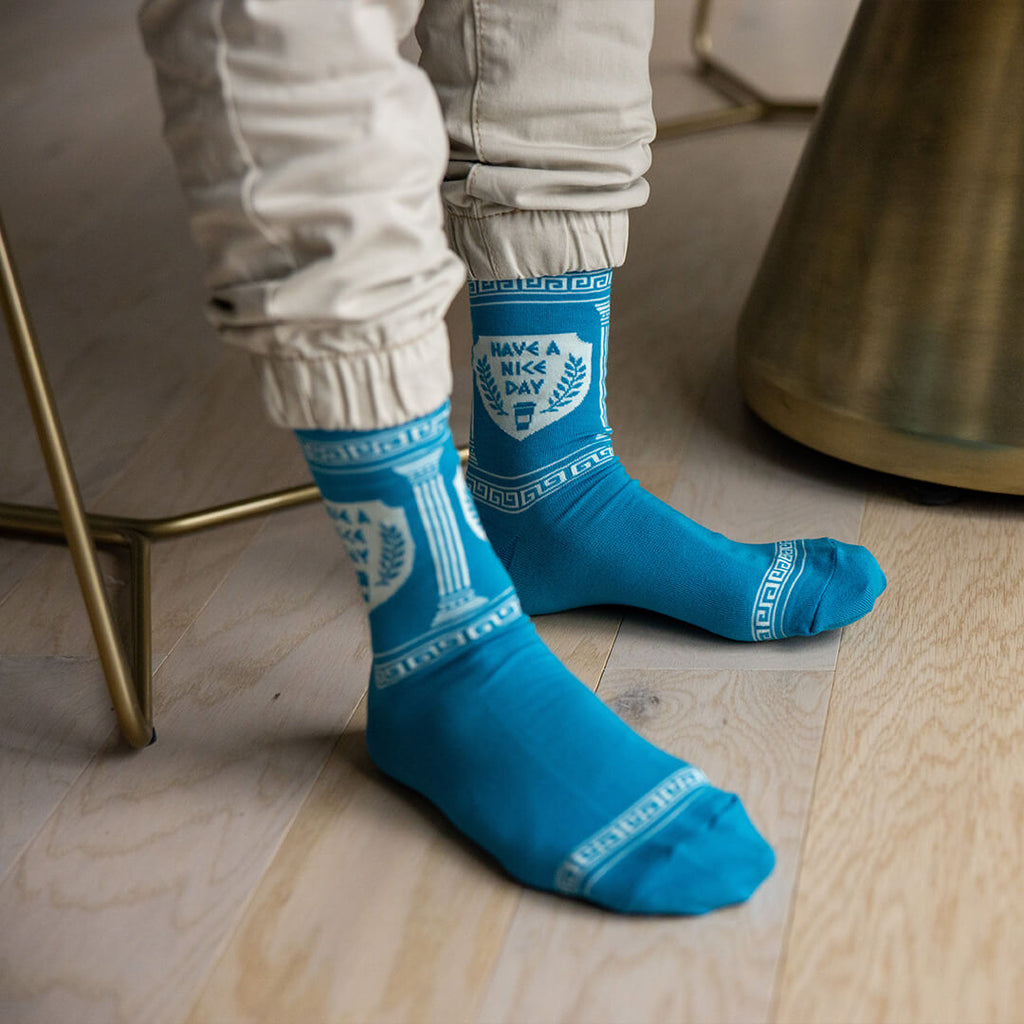 A person wearing bright blue crew socks with white Greek key and classical column patterns, standing on a light wood floor next to a brass chair.