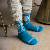 A person wearing bright blue crew socks with white Greek key and classical column patterns, standing on a light wood floor next to a brass chair. thumbnail
