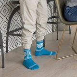 A person wearing bright blue crew socks with white graphic prints and beige jogger pants, standing on a light wood floor in a modern interior with patterned wallpaper and chairs. thumbnail