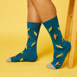 A person's legs wearing blue crew socks with a pattern of cartoon kayaks, standing against a solid yellow background next to a wooden stool. thumbnail