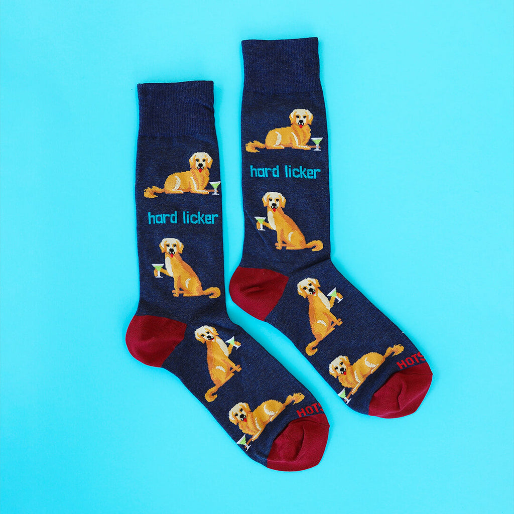 A pair of navy blue crew socks with a repeating pattern of a golden retriever dog holding a cocktail glass and the text 'hard licker', with red heels and toes, displayed on a bright cyan background.