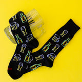 A pair of black novelty socks with embroidered beer bottles and beer mugs, displayed on a bright yellow background next to a clear glass mug.