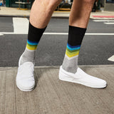 A person wearing gray crew socks with a black top band and multicolor chevron stripes in blue, green, and yellow, paired with white slip-on shoes on a city sidewalk.