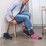 A man sitting on a wooden stool wearing blue jeans, a light blue shirt, and navy blue boots, while trying on a pair of socks with an American flag pattern. The socks feature red and white stripes on one leg and a navy blue background with white stars on the other, with red cuffs. thumbnail