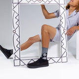A person in light gray pajamas sits behind a large decorative white frame, wearing black slip-on shoes and a gray crew sock with a black-and-white face graphic. The photo is taken in a clean white studio setting.