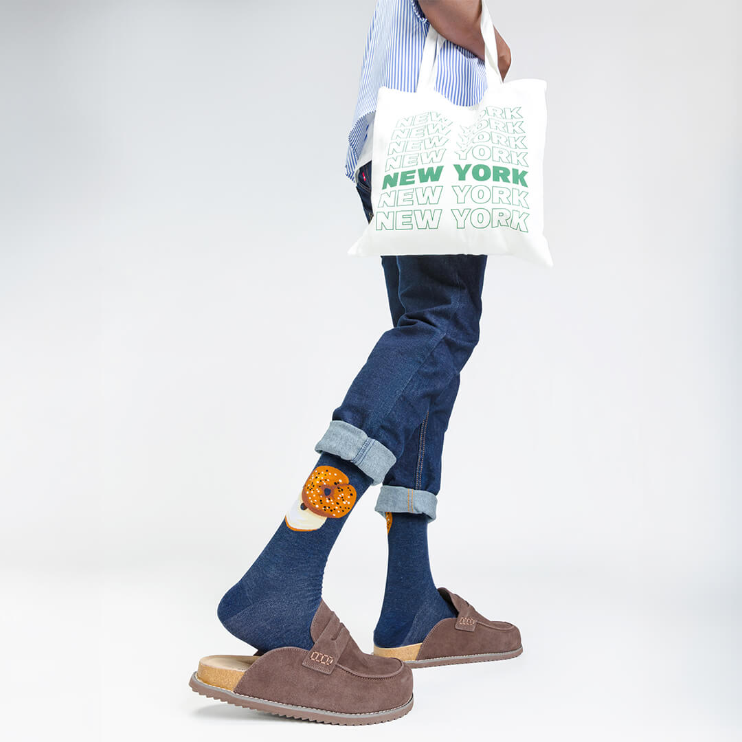 Person wearing blue jeans and brown shoes with a white tote bag featuring 'New York' text.