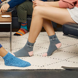 Gray ankle socks with a cat face design worn by a person sitting on a couch in a living room setting, alongside a blue knit slipper and another person's legs. thumbnail