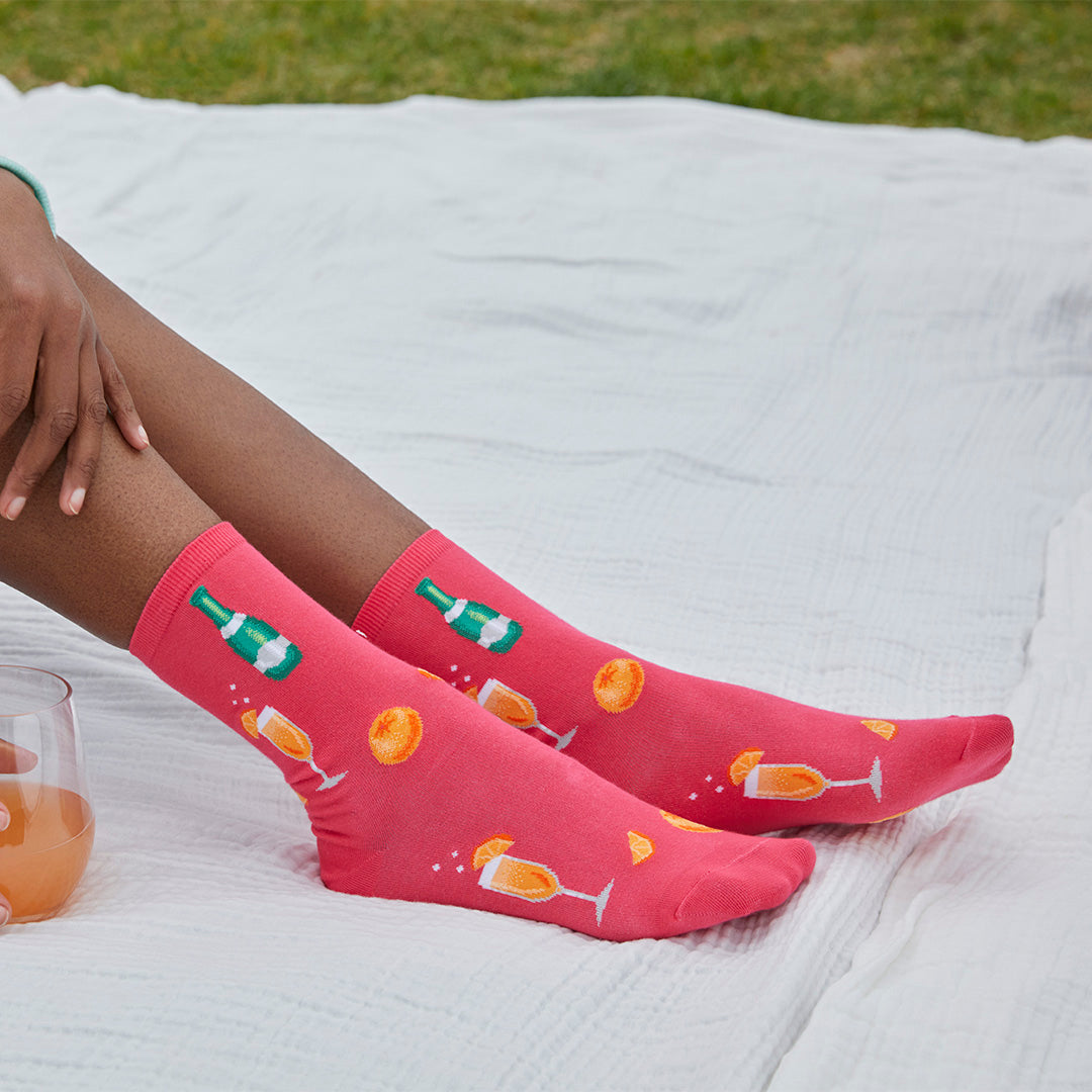A person sitting outdoors on a white blanket, wearing bright pink crew socks with printed champagne bottle, champagne glasses, and orange slices. A glass of orange drink is visible at the lower left.