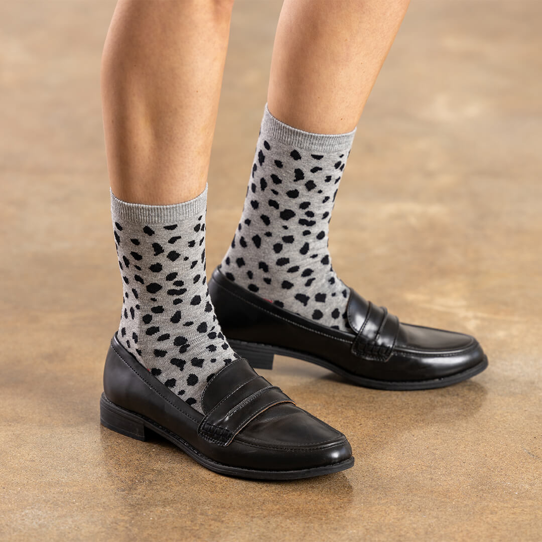 Gray crew socks with a black leopard print pattern, worn with black leather penny loafers on a person's feet against a warm brown floor.