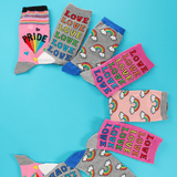 Six pairs of colorful novelty socks arranged in a circle on a bright turquoise background. The socks feature various designs including rainbow clouds, the word 'LOVE' repeated, 'PRIDE' with a rainbow, and blue cuffs. thumbnail