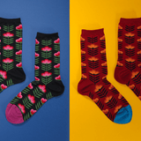 Two pairs of patterned crew socks displayed on a split blue and yellow background. The left pair is black with pink flowers and green leaves, and the right pair is dark red with orange and yellow leaves and blue toes.