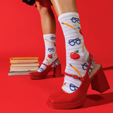 A person wearing white striped socks with red apples, blue glasses, and yellow pencils, paired with red Mary Jane high heels, posed against a solid red background with a stack of books.