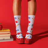 White knee-high socks with a striped pattern and school-themed illustrations of red apples, yellow pencils, and blue glasses, worn with red Mary Jane shoes against a solid red background.