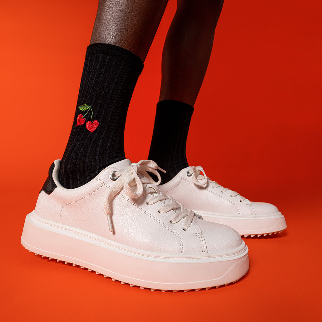 White sneakers worn with black socks featuring red cherries on a red background