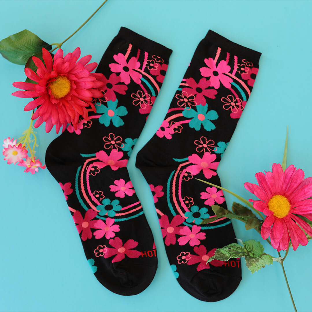 A pair of black crew socks decorated with vibrant pink, teal, and orange floral patterns, arranged on a light blue background with decorative pink flowers.