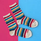 Three crew socks with multicolor horizontal stripes in red, orange, yellow, teal, blue, black, and white, laid out on a split pink and blue background. The toes are cream-colored, and the word 'HOTSOCKS' is printed in red on the toes.