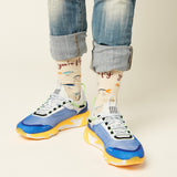 A person wearing cuffed blue jeans, patterned cream socks with text and fish illustrations, and chunky blue, white, and yellow sneakers against a plain light beige studio background. thumbnail