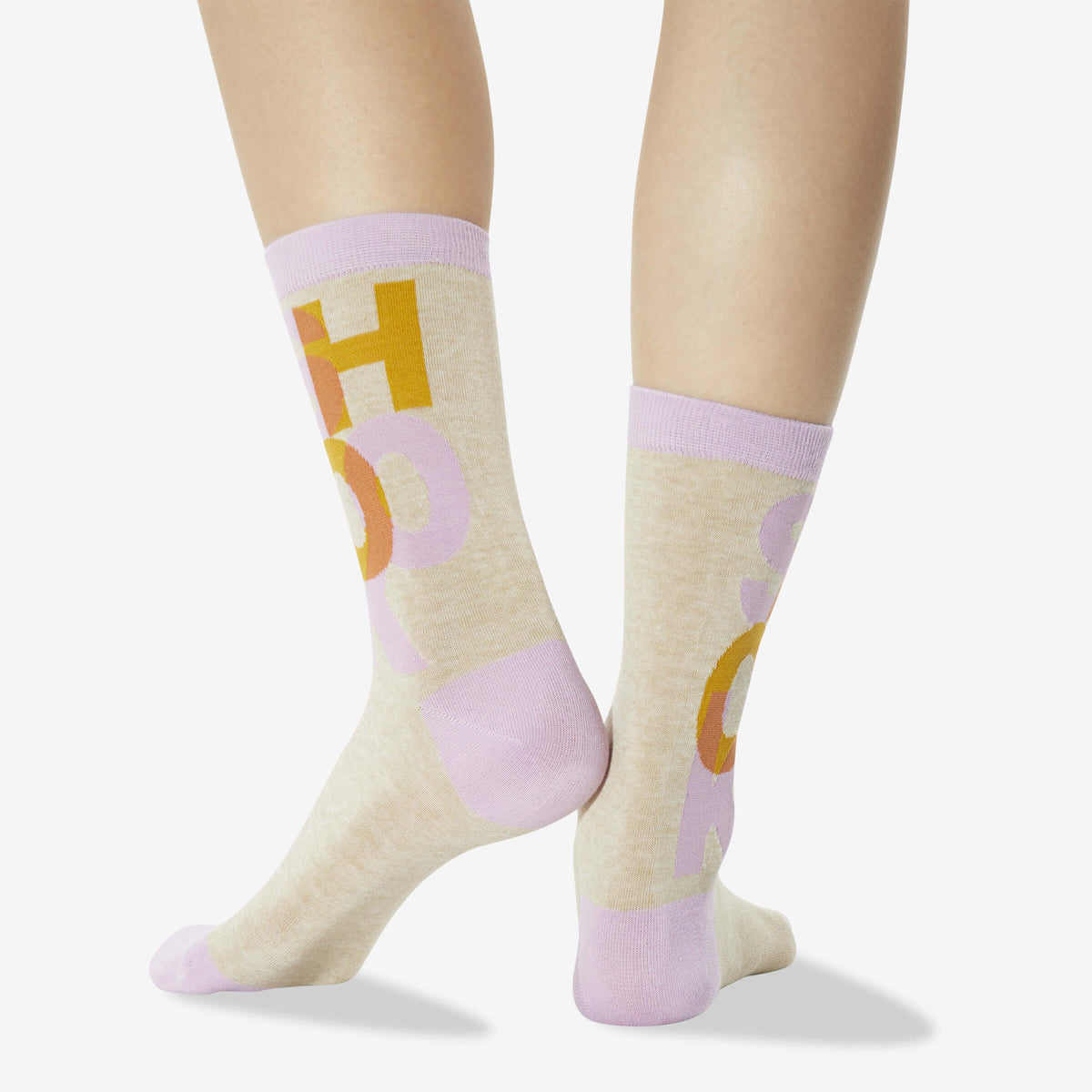 A pair of crew socks worn on a model's legs, featuring a light beige heathered base with lavender cuffs, heels, and toes, and a graphic print of yellow and lavender letters.