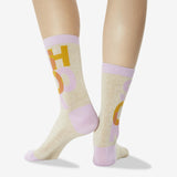 A pair of crew socks worn on a model's legs, featuring a light beige heathered base with lavender cuffs, heels, and toes, and a graphic print of yellow and lavender letters.