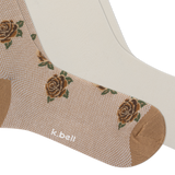 A close-up of a beige sock with a woven rose pattern, brown toe and heel, another pair of light cream socks, and the brand name 'k.bell' printed near the ankle.