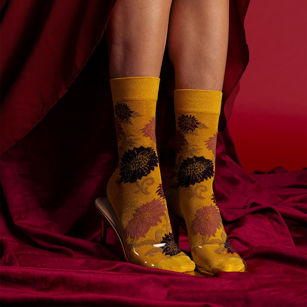 A pair of metallic gold crew socks with a dark floral pattern, worn with clear high-heeled shoes and shown on a model's feet against a draped dark red background.
