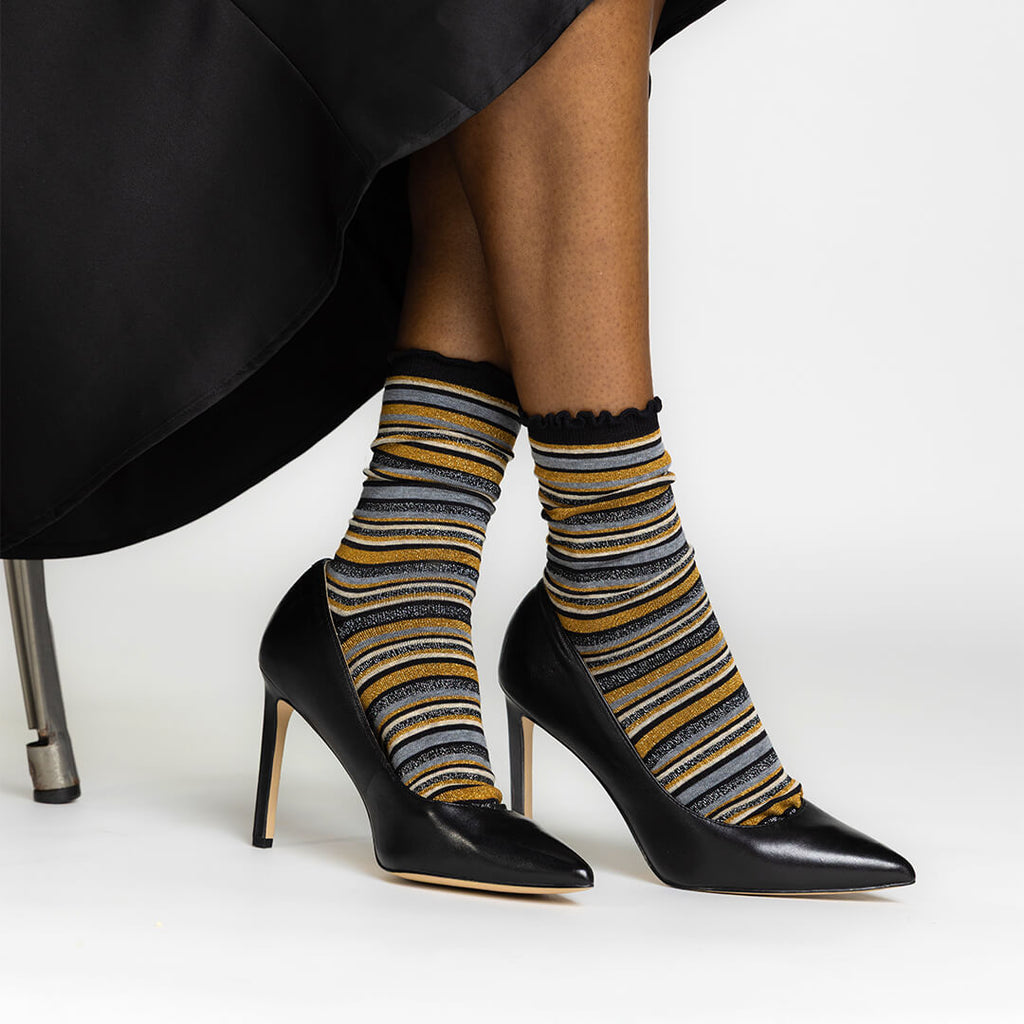 A person seated in a black chair wearing black pointed-toe high heels and metallic-striped crew socks with black ruffled tops. The socks have horizontal stripes in black, mustard yellow, gray, and white.