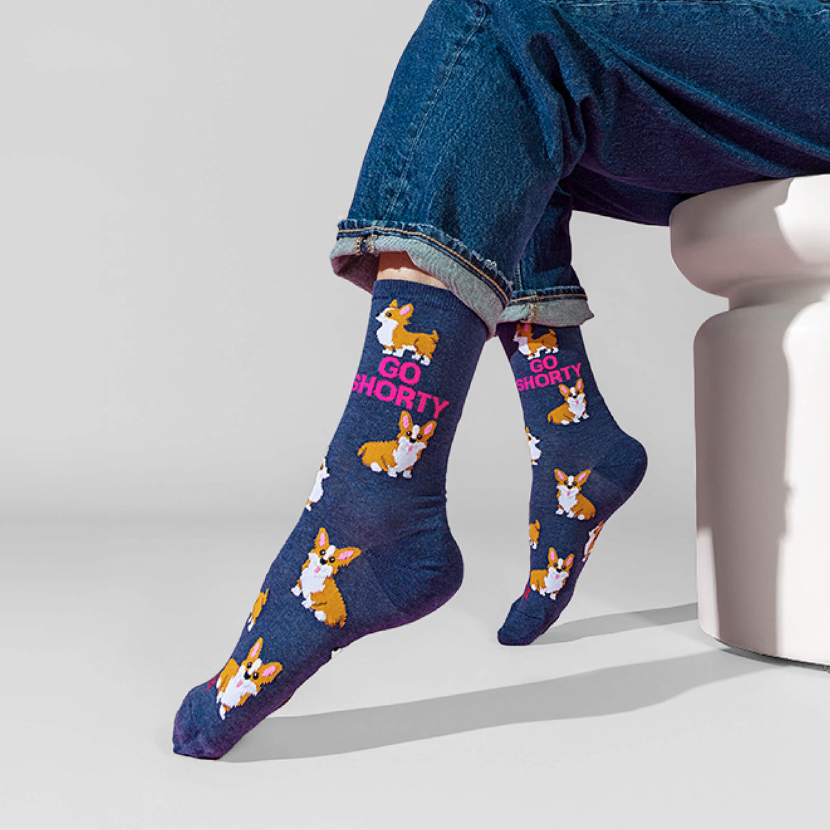 A person seated on a white stool wearing navy crew socks with corgi illustrations and the text 'GO SHORTY' in pink, paired with blue jeans against a light gray studio background.