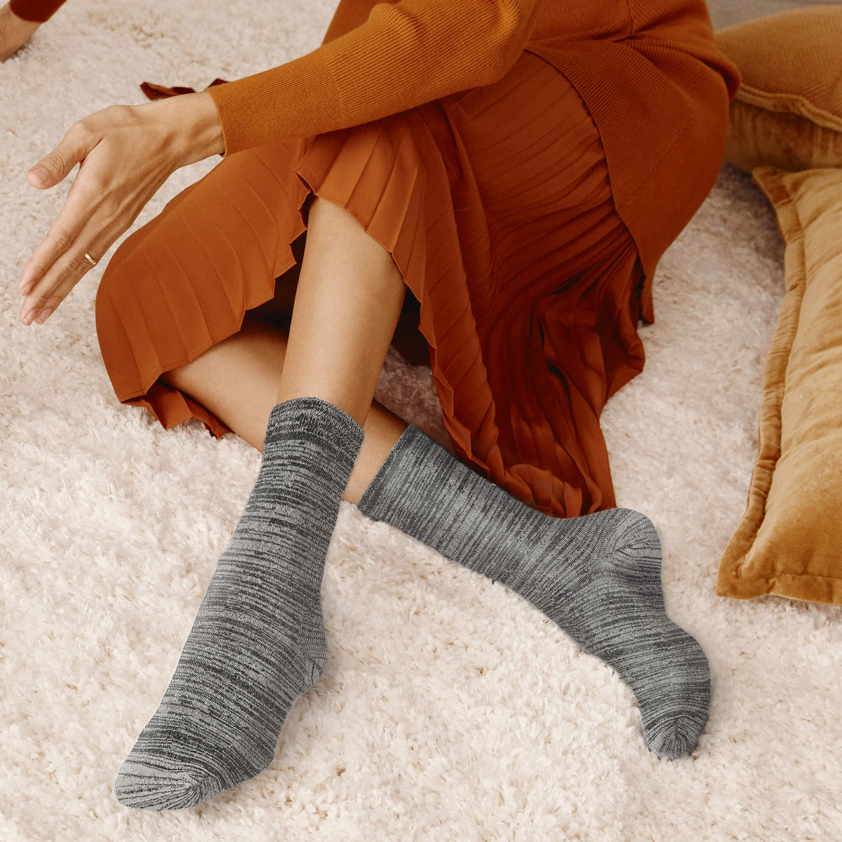 A person seated on a white shag rug wearing gray marled crew socks and an rust-orange pleated dress, with a tan pillow visible at the right edge.